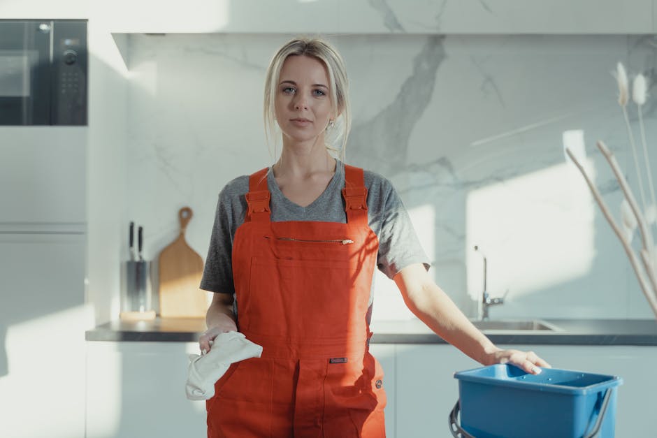 A female cleaner with blonde hair tied back, wearing a grey t-shirt and bright orange apron, stands in a modern kitchen with a white marble backsplash and cabinetry. She holds a white cloth in one hand and is placing her other hand on a blue cleaning bucket positioned on a kitchen counter. The surface appears clean and glossy, with natural light illuminating the area, highlighting the polished marble and stainless steel sink. Visible tools include a wooden cutting board and a knife block in the background, and the overall scene reflects thorough surface cleaning and domestic hygiene, consistent with professional cleaning standards offered by One-Off Cleaning. The setting emphasizes a spotless, well-maintained kitchen environment.