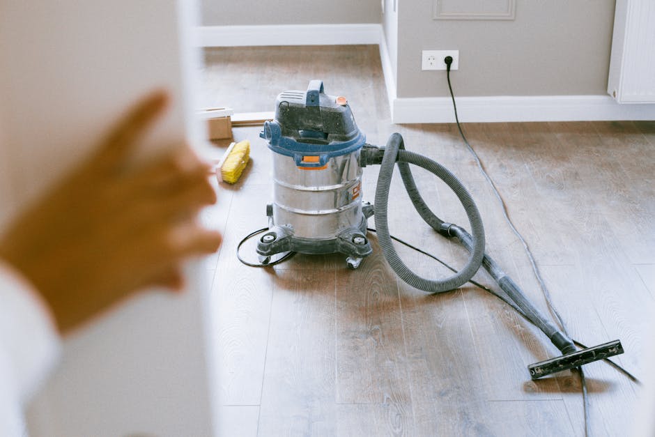 A close-up view of a vacuum cleaner with a metallic canister, black and orange accents, and a flexible black hose attached to a cleaning head, positioned on a wooden floor in a residential living room. The room features a light grey painted wall with an electrical outlet and a white baseboard. Adjacent to the vacuum cleaner, there is a yellow cleaning brush and other cleaning tools on the floor. Soft natural lighting highlights the smooth, dust-free surface of the wooden flooring, reflecting a clean and well-maintained environment, indicating surface cleaning through professional domestic cleaning services by One-Off Cleaning.