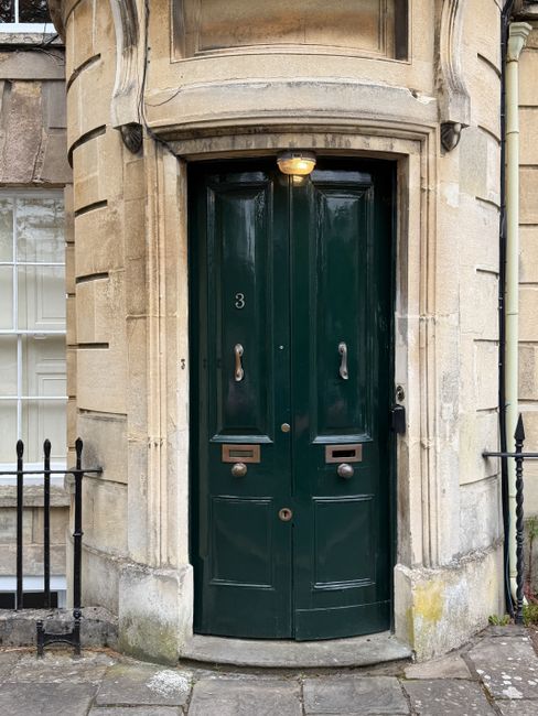 A close-up view of a dark green wooden front door with two vertical panel sections, set within a beige stone building exterior. The door features two silver D-shaped handles positioned at different heights and a letterbox at waist level on each panel. Above the door, a small semi-circular stone canopy is visible, with a warm yellow light fixture illuminating the entrance area. The surrounding stonework shows slight weathering and staining, with a small section of black metal railing on each side of the entrance and a paved stone path leading up to the door. This scene exemplifies surface cleaning and maintenance typical of professional domestic cleaning services provided by One-Off Cleaning, emphasizing the importance of hygiene and visual appeal in residential entrances in the UK.
