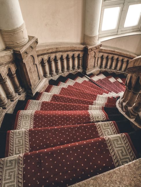 A curved staircase with a red carpet runner featuring a pattern of small white dots and Greek key motifs along the edges, ascending through a historic interior with beige walls and ornate stone balustrades. The staircase is well-maintained and clean, illuminated by natural light coming through large, multi-pane windows at the top. The stone columns and balustrades are polished and free of dust or marks. This setting showcases a classic, elegant space typical of a Victorian-style building. For effective surface cleaning, especially to remove stains from wool carpets, One-Off Cleaning recommends a detailed, professional approach that ensures hygiene and maintenance of such luxurious interiors.